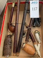 Overview of assorted pipes of various styles and materials in a box, including carved wood, wicker, ceramic, and wooden pipe stems.