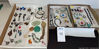 Two cardboard trays containing mixed sterling silver jewelry on white padding. Left tray holds earrings, carved jade pieces, pendants and bracelets; right holds additional earrings, bracelets and necklaces.