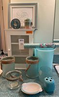 White wooden cabinet with glass front, glass cylinder candle holders with rope handles, ceramic soap holder with shell design, unused candle, geode slice clock with gold hands on top, framed shell print hung behind, teal plastic wastebasket, teal ceramic toothbrush holder, and teal glass candle bowl arranged on a teal countertop.