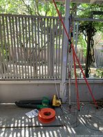 Two leaf blowers, one yellow and one green, with an orange cord reel on floor against porch railing. Red handled extension branch cutters leaning against wall.