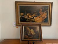 Two framed art pieces hung and displayed together. Larger piece with musical instruments, smaller showing a man teaching a child guitar.