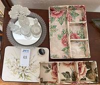 Photo showing a round silver tray holding three clear glass bottles and two votive holders. Next to the tray is a table protector with floral design and two floral cloth-covered storage boxes.
