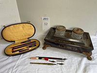 Wide angle view showing black-lined case with dip pens and nibs alongside the Regency brass inlaid ink stand with glass ink wells and wooden lids.