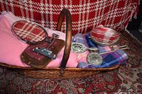 Wicker basket filled with pink and purple plaid blankets, two handheld round mirrors, and a silver-toned brush and hand mirror set.