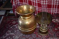 Photo of brass spittoon and brass oil lamp placed on patterned carpet in front of red and white plaid upholstery.