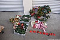 Wide view of lot of assorted Christmas wreaths, small tree, candy cane bows, Merry Christmas sign, and more decorations on concrete floor in front of metal door.