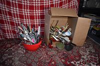 Overview of red plastic bowl filled with various metal and plastic kitchen utensils including knives and spatulas, next to cardboard box containing more utensils such as ladles and strainers.