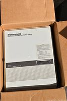 Panasonic phone switch unit inside a cardboard box, white plastic body with labels and vents, appears new and unused.