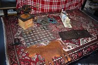 Wide view of all items laid out on a patterned red and white couch with a rug beneath showing vintage grinders, a metal butter imprint pan with cow designs, a rust-colored cow-shaped trivet, and other miscellaneous kitchen tools.