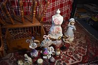 Assorted vintage perfume bottles and figurines displayed on carpet in front of red and white blanket backdrop.