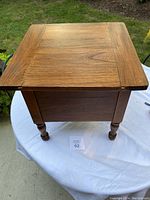 Front angled view of the wooden antique commode table, showing the top with wood grain and turned legs, placed on a white cloth outside.