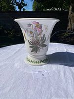 Full view of the white ceramic cache pot on white cloth with outdoor background showing botanical and butterfly motifs.