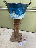 Blue metal Christmas bucket on wood stand featuring snowman and Christmas tree winter scene with Merry Christmas sign