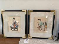 Two framed Chinese watercolour paintings side by side on a table, showing female figures in traditional attire, gold and black frames with mats