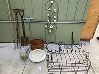 Photo of garden tools, metal flower trellis, wicker basket, glass cloche, plastic plant saucer, and large metal window box planter on concrete floor