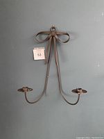 Metal wall hanging candle holder shaped like a bow with two candle holders, attached to a gray wall with a numbered tag visible.