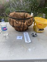 Wide view showing the hanging basket with coconut coir liner, yellow Vidor hand spreader, white wall thermometer, and metal hose splitter on a concrete surface outdoors.