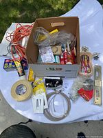 Top-down photo of the box with visible extension cord, masking tape, various caulk tubes, small hardware packages, and electrical parts