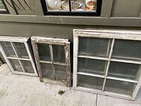 Six vintage wooden window panes leaning against a green wall outdoors showing frame details and glass intact.