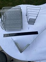 Photo showing the three metal mesh letter trays stacked, the vertical folder rack, the black magnetic strip for wall mounting, and the silver metal wall shelf support bracket all placed on a white surface.