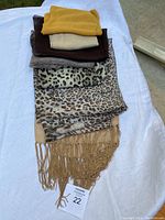 Stack of 7 scarves in varying brown, beige, gold, and animal print colors on a white cloth background; includes fringing on one scarf.