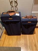 Two upright navy Samsonite suitcases with brown leather handles and trim shown from front on hardwood floor.