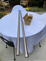 Two extra long brushed nickel metal curtain rods displayed on a white cloth-covered table outside, showing the smooth cylindrical shape and the adjustable feature.