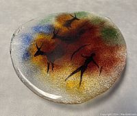 Photo showing the oval glass dish with colorful Indigenous-style painted figures on a textured silver background.