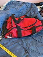 Side view showing red fabric, black mesh panels, black handles, Adidas logo on side, with measuring tape alongside for scale.