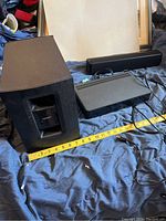 Photo showing Bose subwoofer, center speaker, and control center on a dark fabric surface with measuring tape in front, illustrating size and condition.