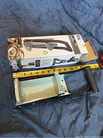 Photo of the BK can crusher with ruler measuring its length approximately 15 inches, placed on a blue surface with packaging box showing the product name and recycling graphics.