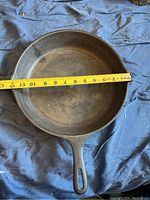 Top view showing 12 inch diameter pan with measuring tape across the cooking surface, black cast iron frying pan with handle on blue cloth background.