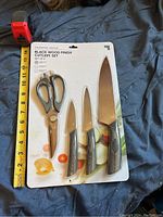 Front view of sealed Sharper Image cutlery set including three knives and scissors with black wood finish handles, next to measuring tape for size reference