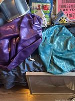 Photo showing two partially deflated exercise balls, one purple and one teal, placed on a black fabric surface with various background items like a plant bag and storage bin.