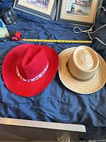 Two hats side by side on a dark fabric surface showing a red fabric fedora with Canadian flag band and a beige woven straw fedora with brown band.