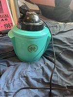 Front view of the humidifier showing the Sunbeam logo on the turquoise body and black top dome with power cord attached.
