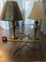 Two similar table lamps on hardwood floor with measuring tape in front showing approximately 19-inch height. One lamp has beige fabric shade; the other has dark shade wrapped in plastic.