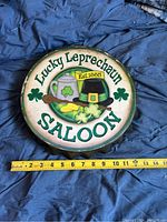 Front view of round metal sign with 'Lucky Leprechaun Saloon Est. 1885' text and decorative elements including shamrocks, mug, pipe, pot of gold, showing condition and size scale.