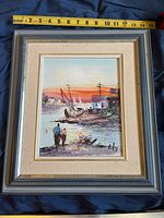 Framed canvas painting showing a harbor scene with boats and two people by the shore at sunset, with a ruler for scale.