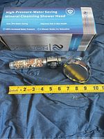 Photo of Nortex high-pressure water saving mineral cleansing shower head with multi-colored mineral beads inside clear handle and chrome shower head on dark fabric background with measuring tape showing size.