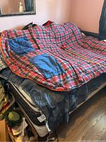 Sleeping bag opened on a queen bed showing plaid pattern with blue patches and orange trim edges.