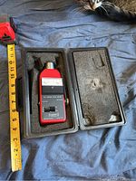 Red Quest Electronics 214 Sound Level Meter in open black foam-lined carrying case next to a yellow measuring tape showing the length around 7.5 inches. A tabby cat is partially visible in the top right corner.