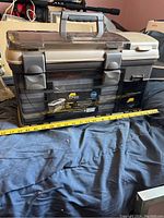 Full view of the closed fishing tackle box showing the Plano label, featuring dual latch locks and a carrying handle with measuring tape for size reference.