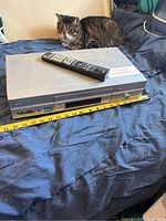 Front angled view of the silver Sony VCR sitting on a blue cloth with a remote control placed on top, a cat nearby for scale.