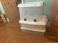 Stack of three clear plastic storage bins with lids. Two larger bins stacked on the bottom, one smaller bin with wheels on top.