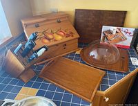 Photo showing a painted wooden breadbox, large butcher block board, wooden charcuterie board with clear dome cover, a small rectangular cutting board, and a wooden knife block holding knives and a wine bottle.
