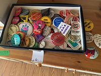 Top view of a box filled with numerous round buttons and badges featuring various feminist and social justice messages, showing diversity of colors and slogans within the lot.