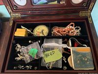 Open wooden jewelry box showing two shelves of assorted costume jewelry, including beads, metal and rhinestone pieces, and a floral round compact.