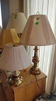 Two brass-colored table lamps with pleated fabric shades side by side on a wooden surface in front of window blinds. Lamps are untested, one taller and one shorter. Lamps have visible power cords and green price stickers.