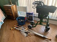 Overview of medical equipment lot including Handike Kpress walker, aluminum crutches, blue bucket with step stool, blood pressure monitor box and other items.